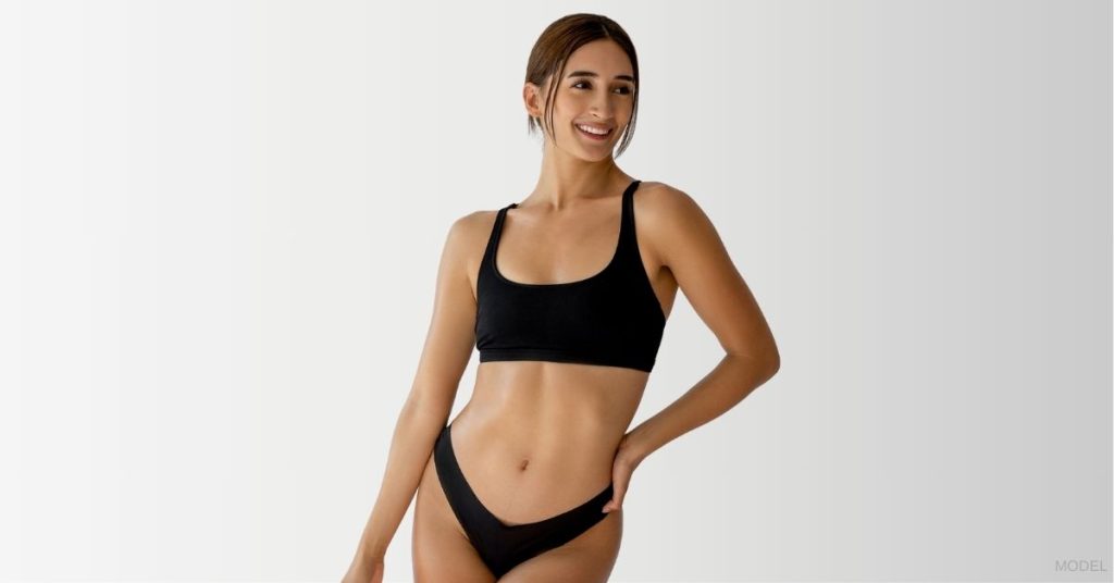 A woman in a black bikini top and underwear with her hand on her hips. (Model)